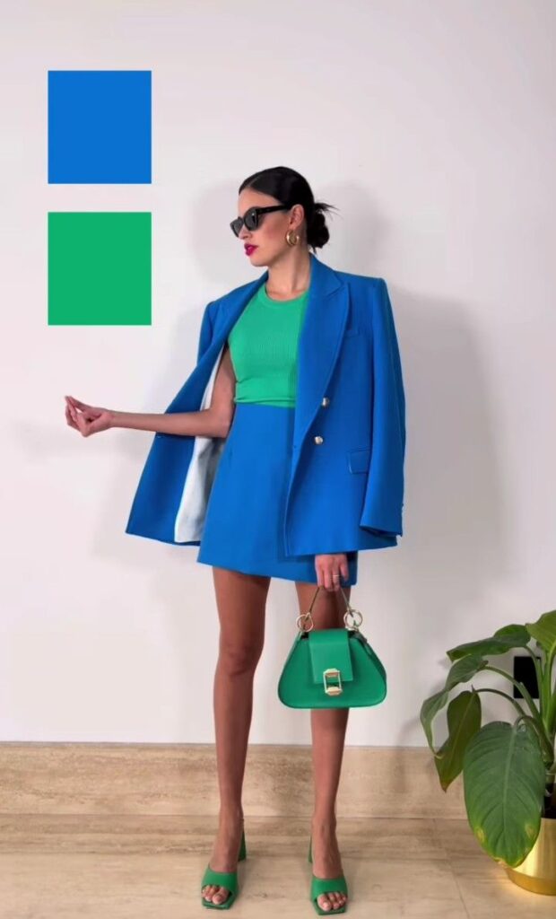 Stylish woman in blue blazer and skirt with green top, heels, and handbag, posing chicly indoors near a plant.