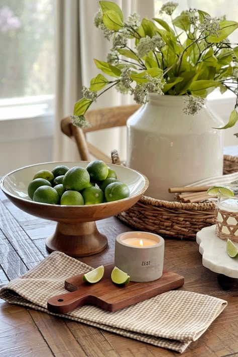 Bowl of fresh limes and lit candle on wooden table setting with flowers in vase, offering a cozy, natural atmosphere.