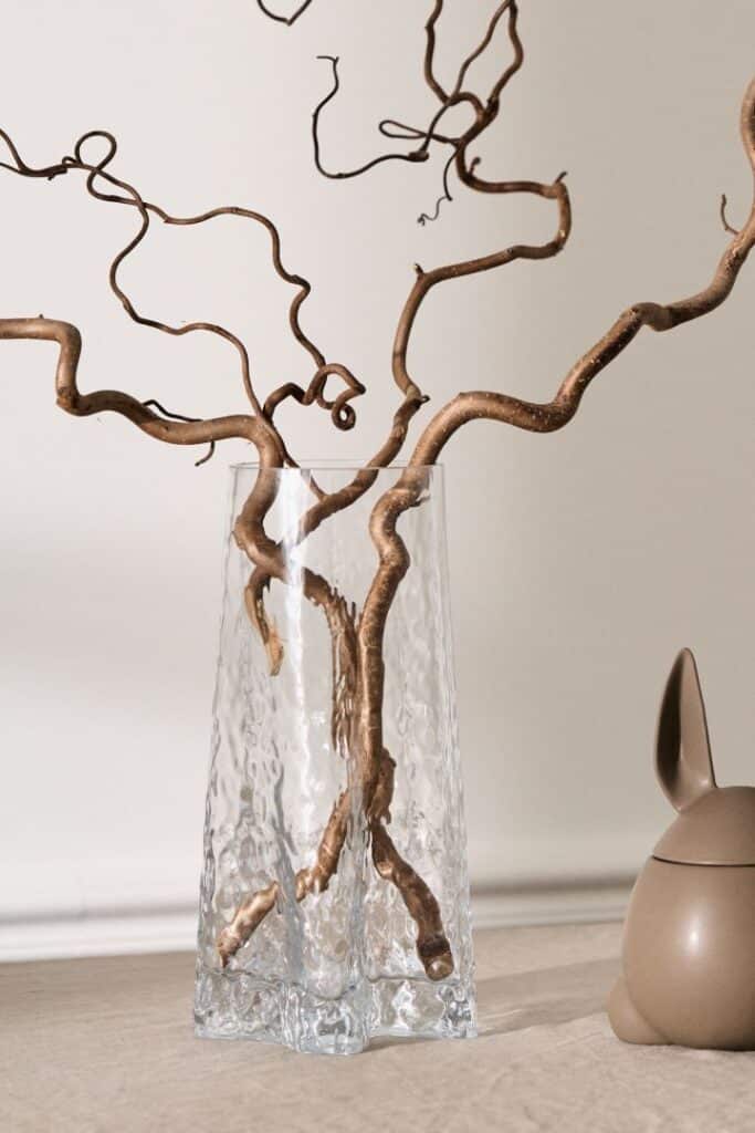 Curly branches in a textured glass vase beside a ceramic rabbit figurine on a beige table. Minimalist decor.