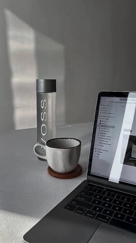 Minimalist workspace with laptop, coffee mug, and water bottle on a white desk, sunlit ambiance.