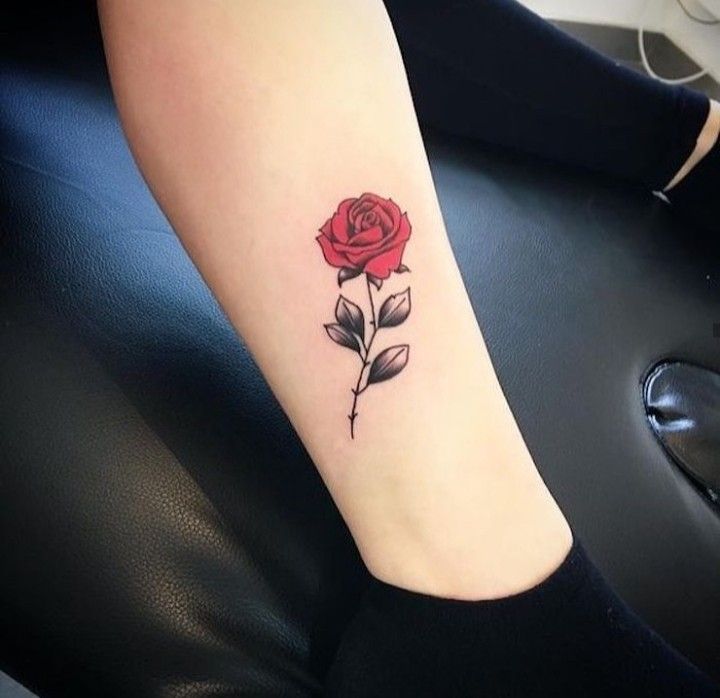 Red rose tattoo on a person's forearm with black leaves on a leather chair background.