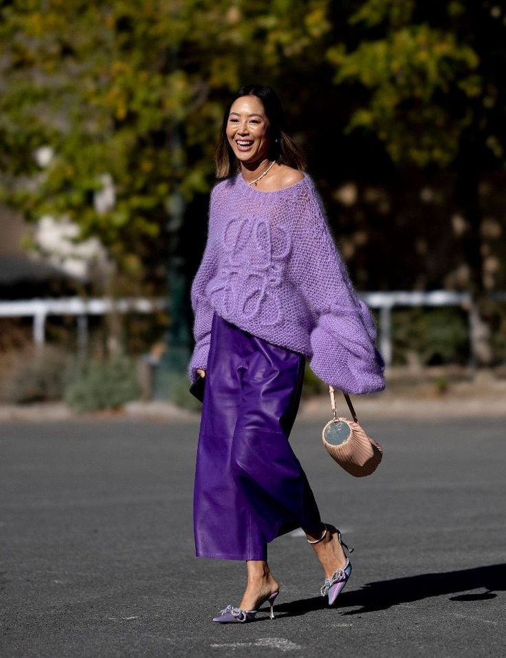 Woman in stylish purple outfit with a knit sweater and leather skirt walking outdoors, carrying a chic handbag.