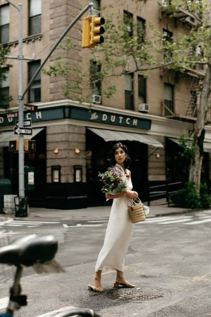 Woman in white dress crossing street, holding flowers in urban setting near The Dutch restaurant. Street style.