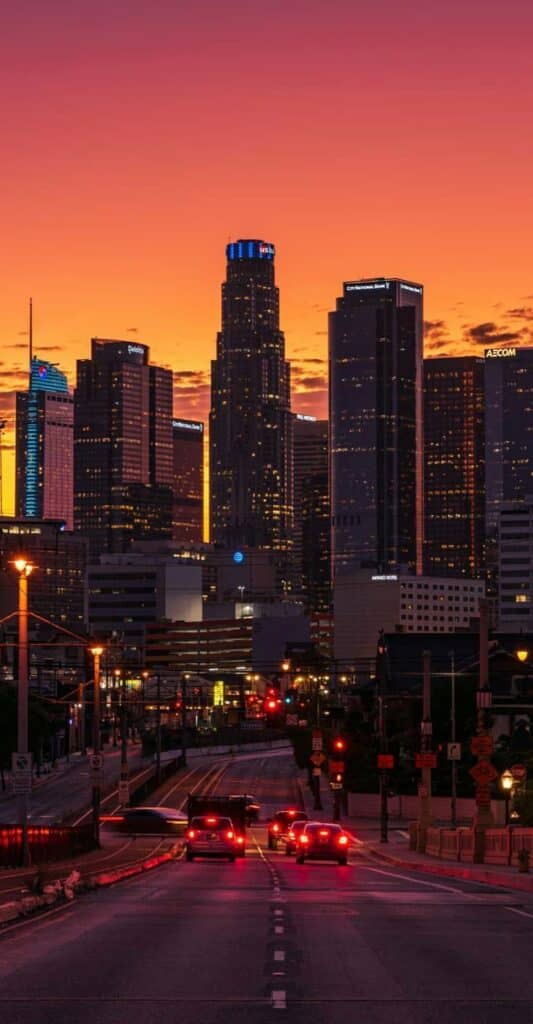City skyline at sunset with cars on the road, vibrant orange sky and illuminated skyscrapers in the background.