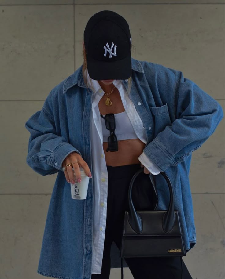 Stylish person in denim jacket, black cap, and crop top holding coffee cup and black handbag, casual streetwear fashion.
