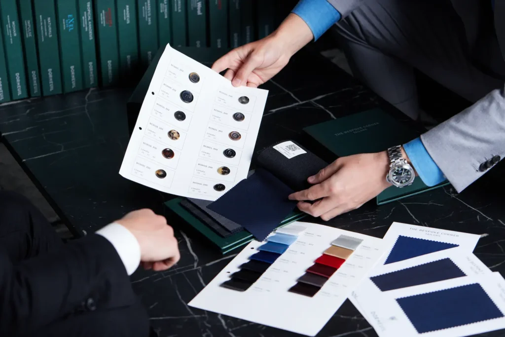 Two people examining fabric swatches and button samples on a table for tailoring decisions.
