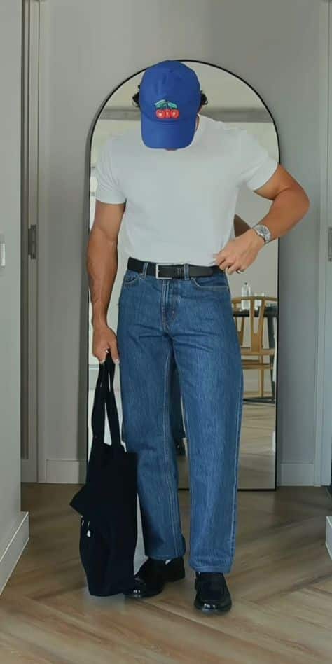 Man in casual outfit with blue cap, white shirt, and jeans holds a black bag in a modern, minimalistic setting.