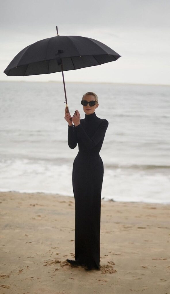 Person in a long black dress holding an umbrella on a beach, wearing sunglasses, with the ocean in the background.