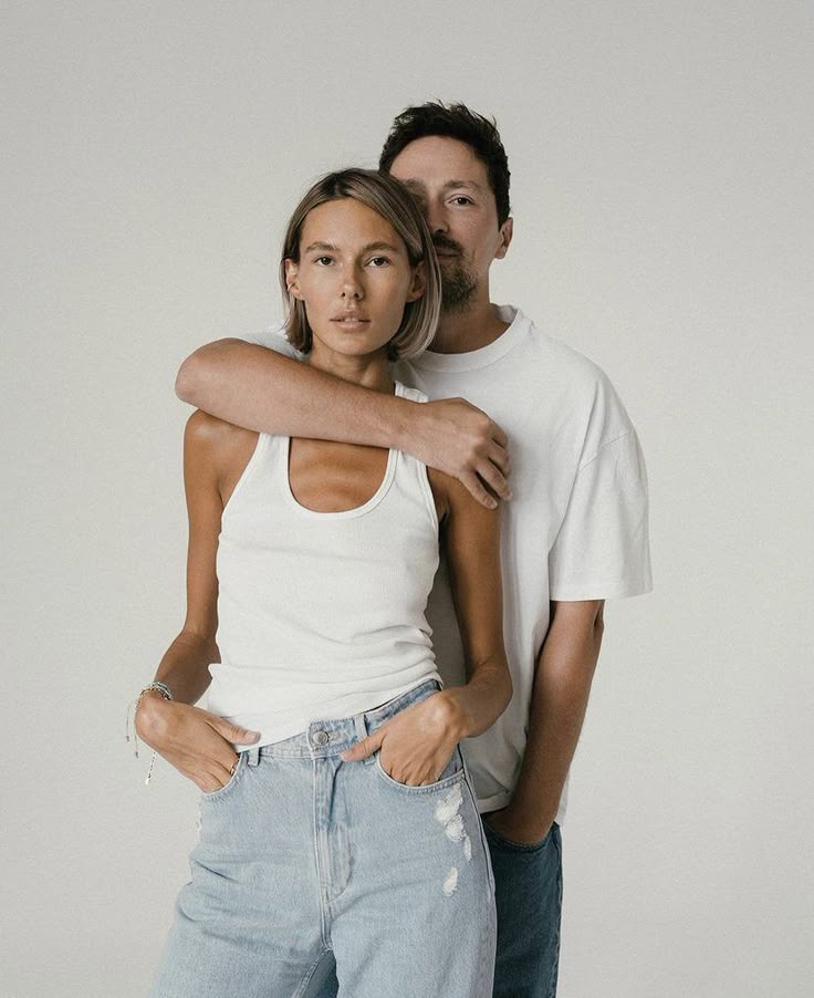 Casual couple posing in neutral tones, with the woman in front, wearing a white tank top and jeans.