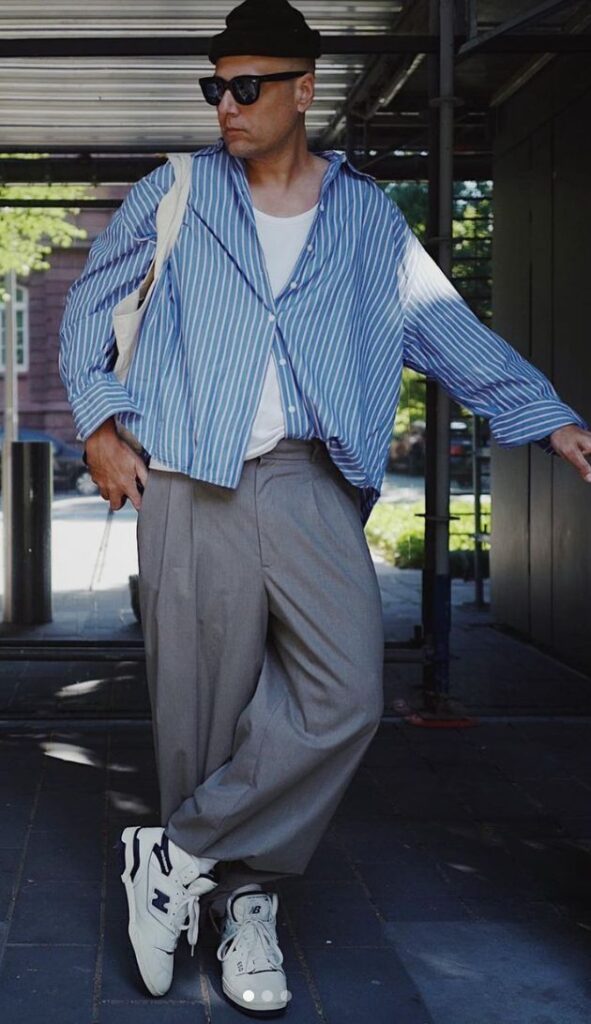 Man in stylish streetwear: blue striped shirt, gray pants, white sneakers, sunglasses, and hat in urban setting.