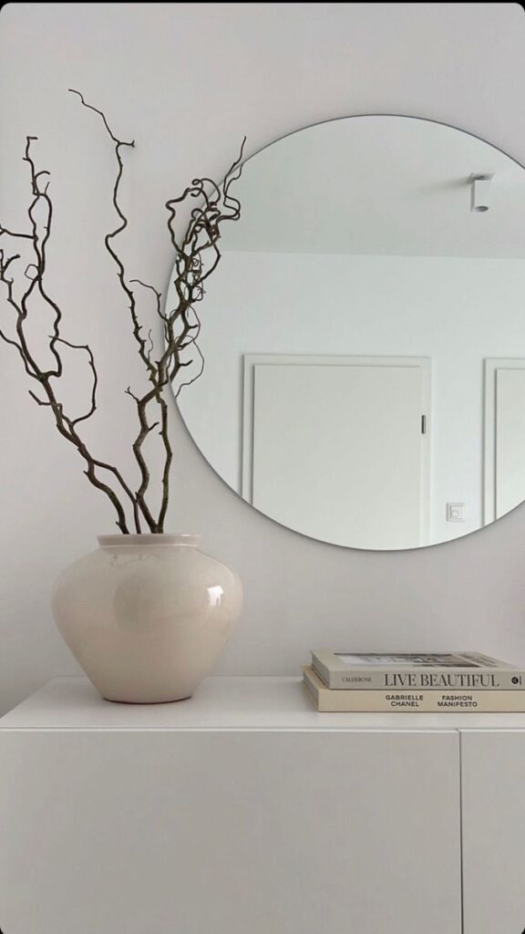 Minimalist decor with a round mirror, vase with branches, and stacked books on a white cabinet.