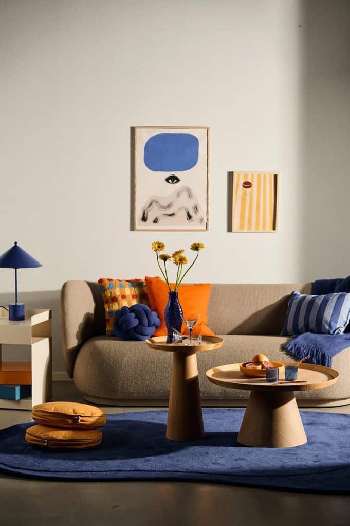 Modern living room with beige sofa, colorful cushions, round tables, abstract art, and a blue rug.