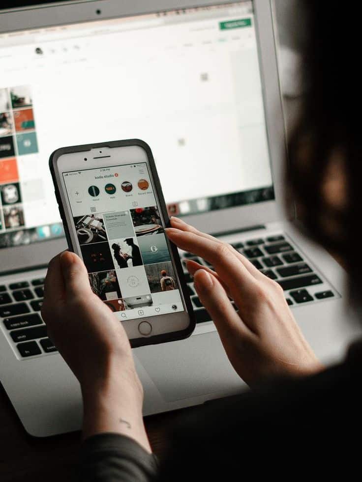 Person browsing Instagram on smartphone in front of a laptop, focusing on social media engagement.