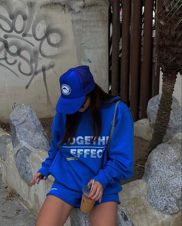 Person in blue outfit and cap holding iced coffee, sitting by urban graffiti wall and rocks. Casual streetwear fashion.