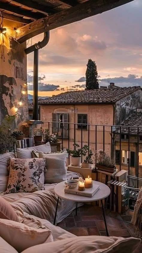 Cozy balcony at sunset with pillows, candles, and plants, overlooking rustic rooftops. Perfect for relaxation and ambiance.