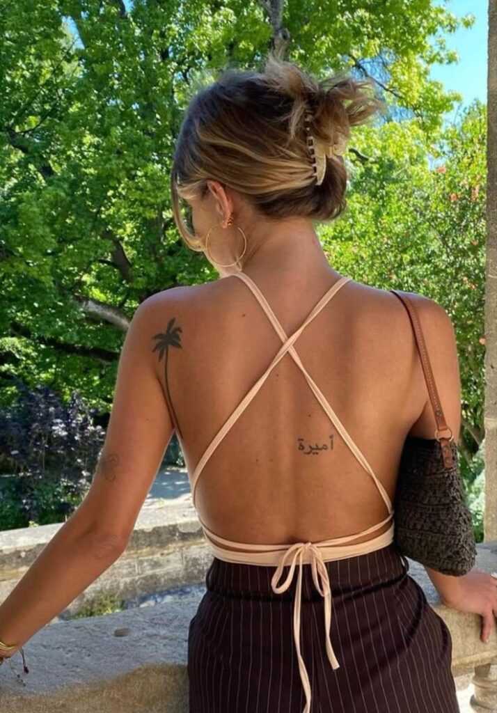 Woman with back tattoo in strappy top and hoop earrings, standing outdoors amidst green trees.