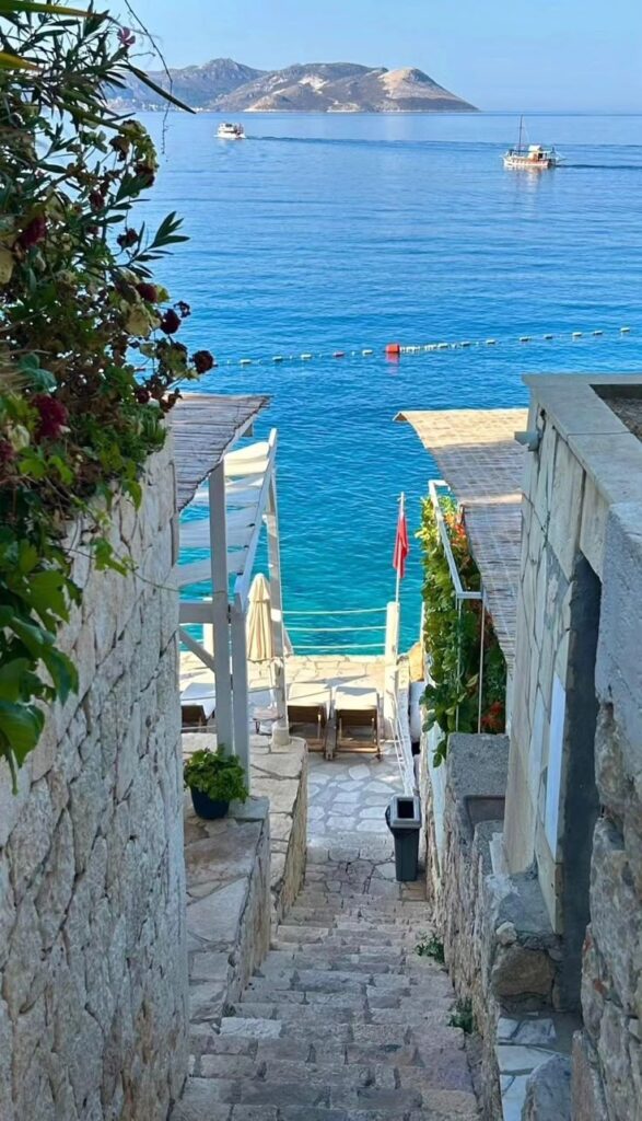 Antalya 2 Stone steps lead to a serene turquoise sea, framed by rustic buildings and distant mountains. | Sky Rye Design Stone steps lead to a serene turquoise sea, framed by rustic buildings and distant mountains.