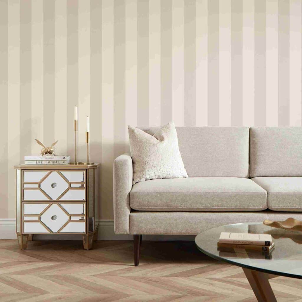 Elegant beige living room with modern sofa, striped wallpaper, mirrored side table, and wooden centerpiece on glass table.
