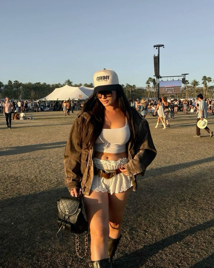 Woman in festival attire with cowboy hat and sunglasses, walking outdoors at an event with tents and crowd in background.