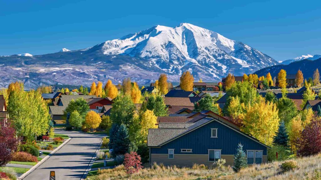 Vibrant autumn foliage and suburban houses with snow-capped mountains in the background under a clear blue sky.