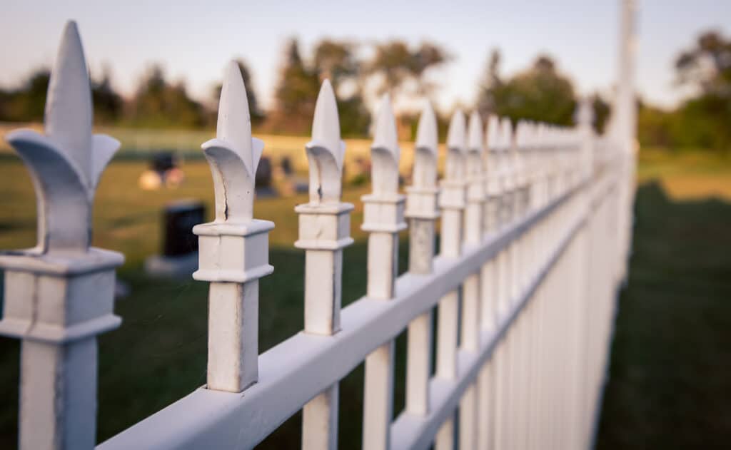 Close-up of a white metal fence with pointed tops, outdoors, blurred background of a green field and trees.