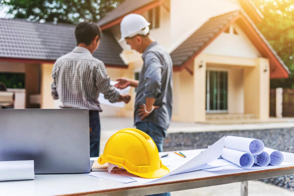 Construction site meeting with two men discussing plans; laptop and hard hat on table in foreground.