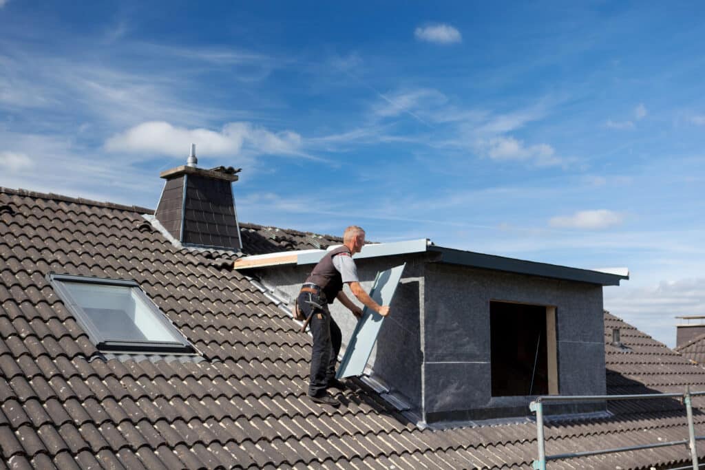 Roofer installing dormer siding on a tiled roof under a clear blue sky.