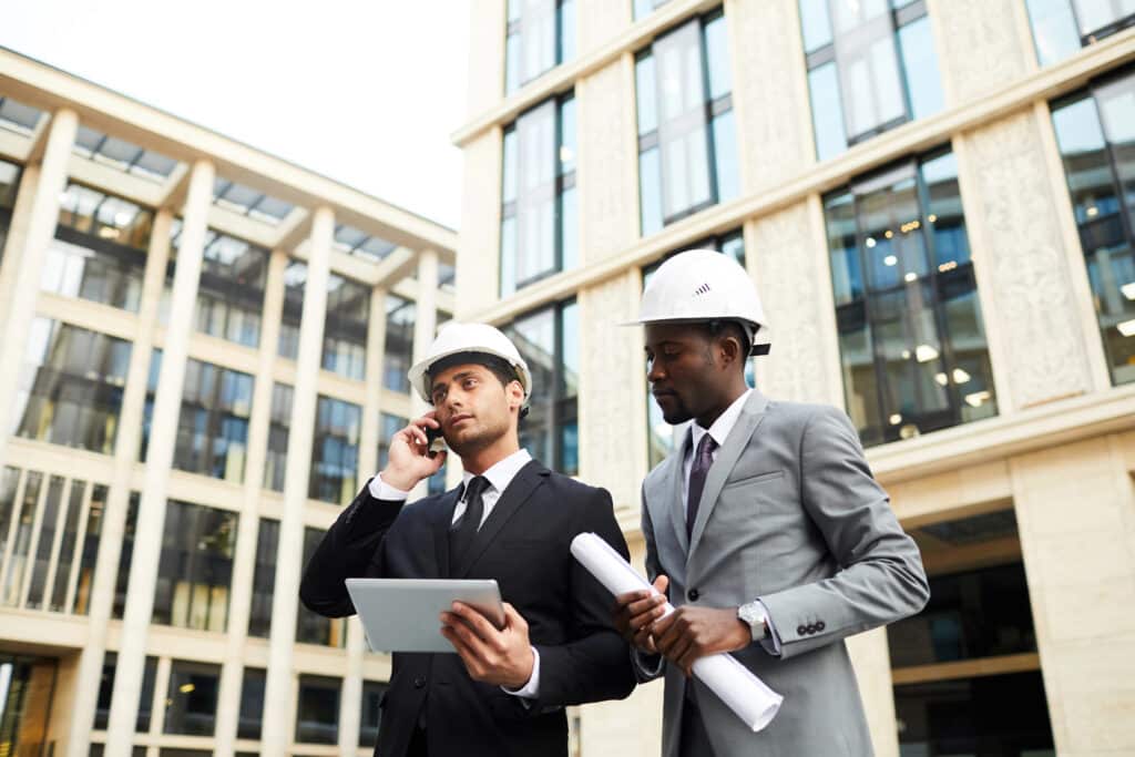 Two engineers in hard hats review building plans and discuss via phone outside a modern office building.