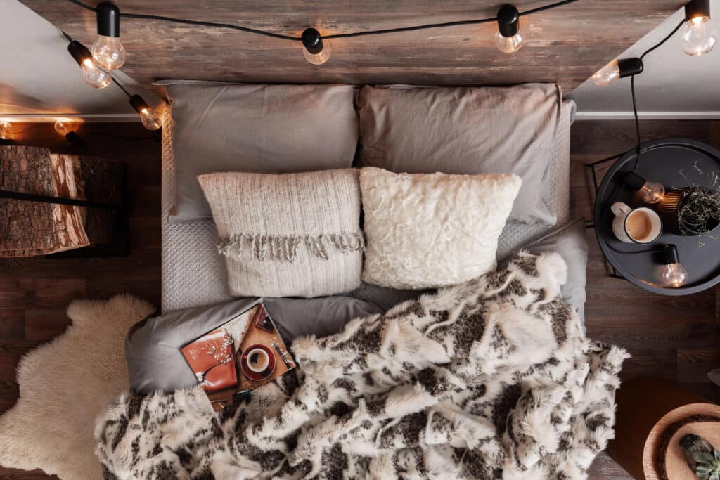 Cozy bedroom scene with fluffy pillows, faux fur blanket, string lights, and coffee on a small round table.