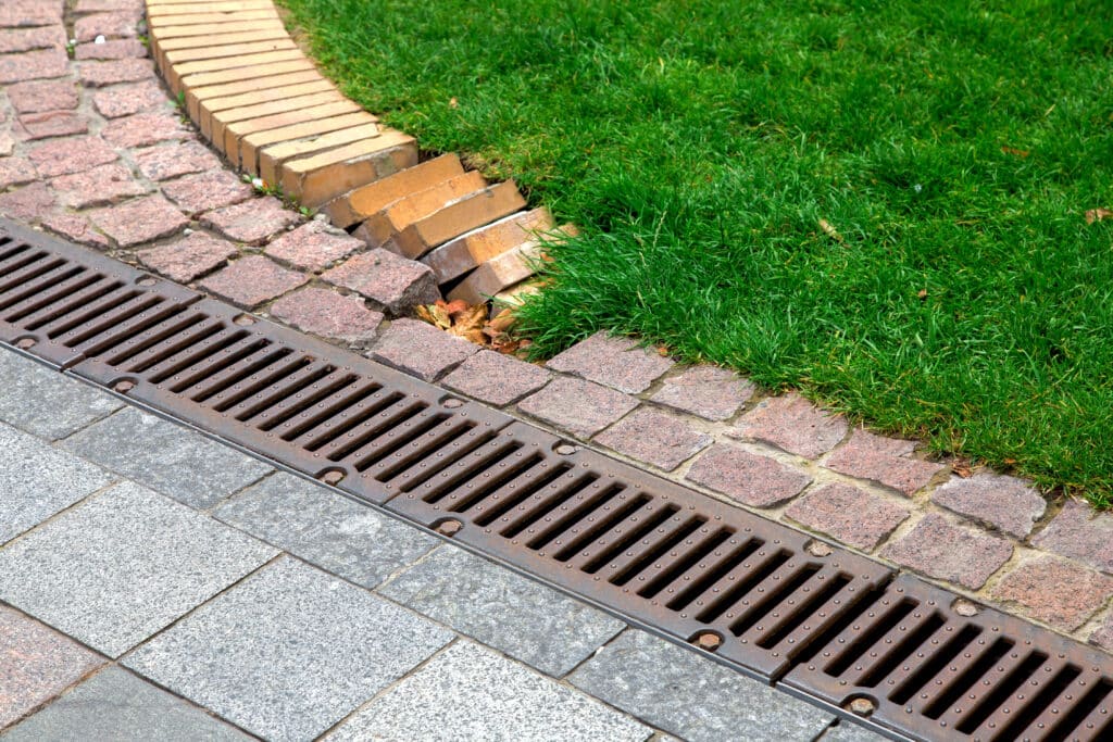 Sidewalk drain with stone bricks and green grass edge, showcasing drainage and landscape design.