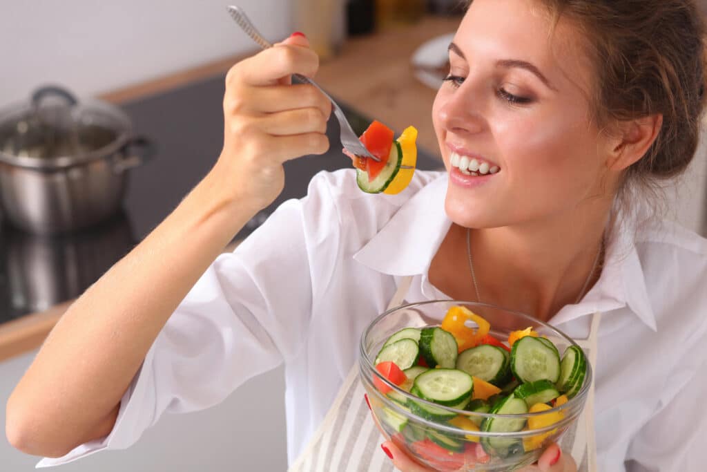 Woman eating fresh vegetable salad from a bowl, smiling in a kitchen setting. Healthy lifestyle concept.