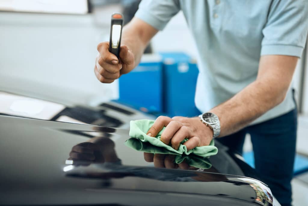 Person inspecting and polishing a car hood with a flashlight and green cloth for quality maintenance.