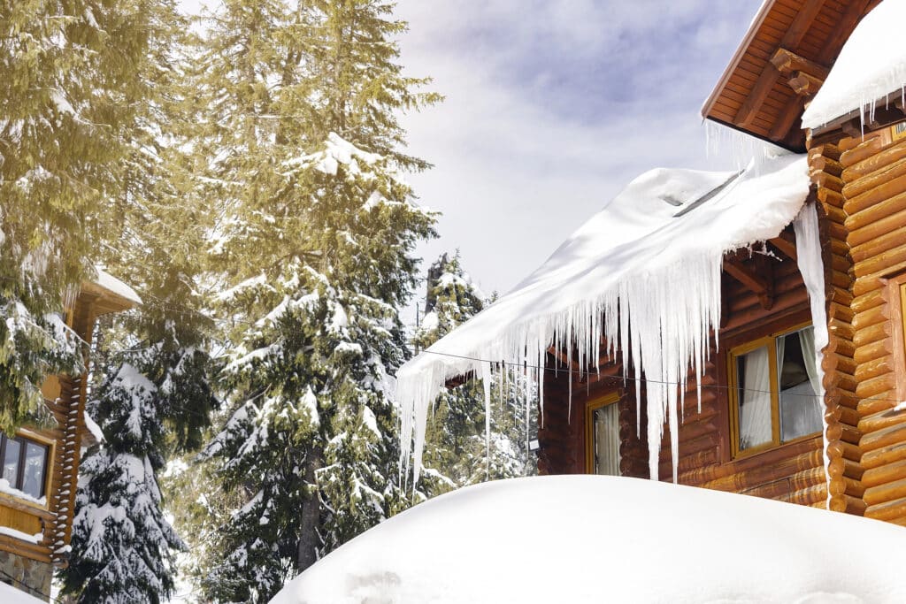 Snow-covered cabin with icicles hanging from the roof, surrounded by snowy pine trees under a clear sky.