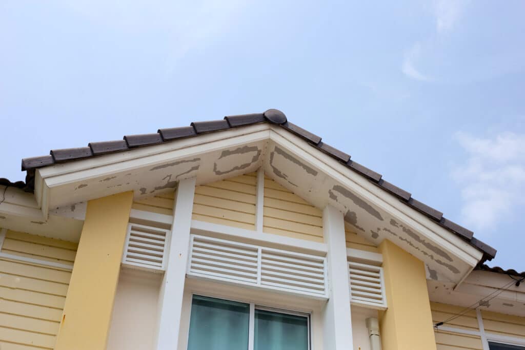 Peeling paint on yellow house gable roof under blue sky highlights need for maintenance and renovation.