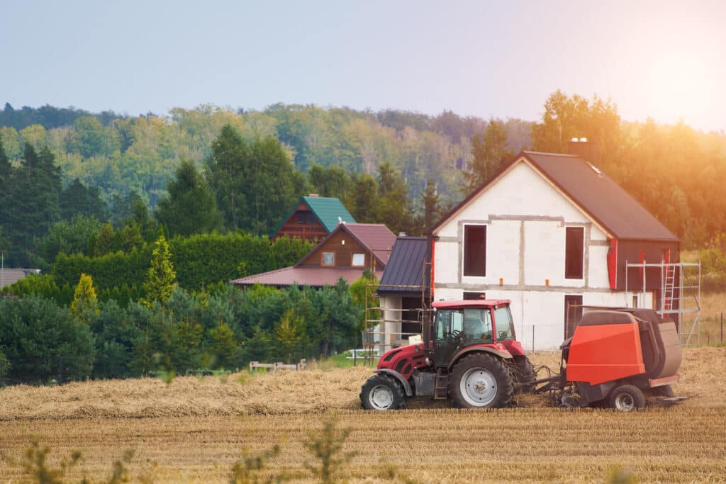 Red tractor baling hay near a country house with lush green trees in the background.