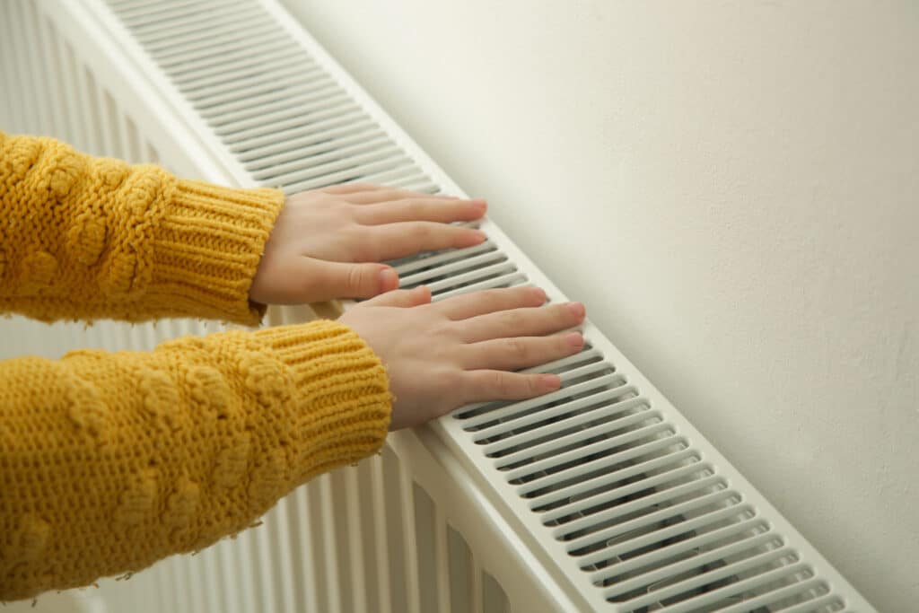 Hands touching a radiator for warmth, wearing a cozy yellow sweater.