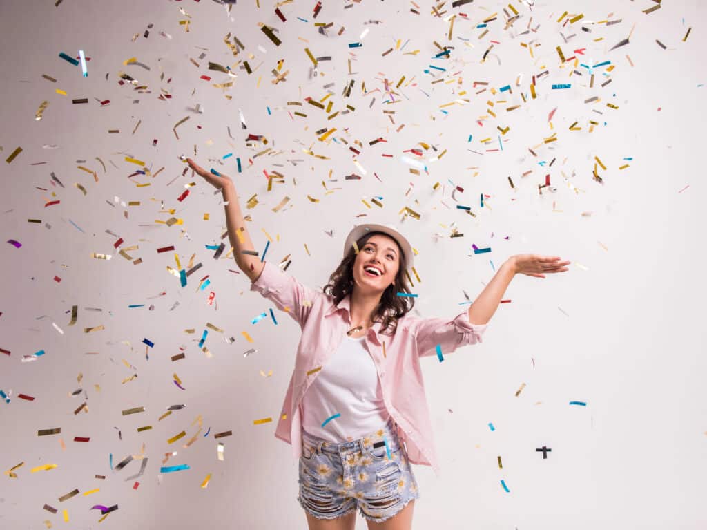 Smiling woman in casual outfit enjoying colorful confetti falling, expressing happiness and celebration indoors.