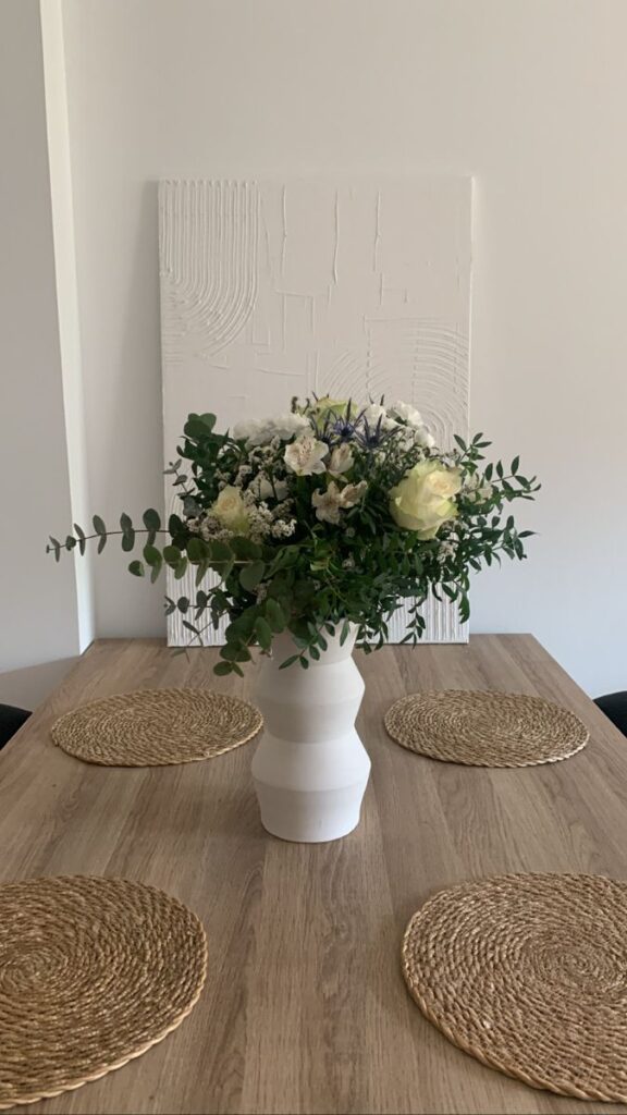Floral arrangement in white vase on wooden table with woven placemats, minimal decor in modern dining room.