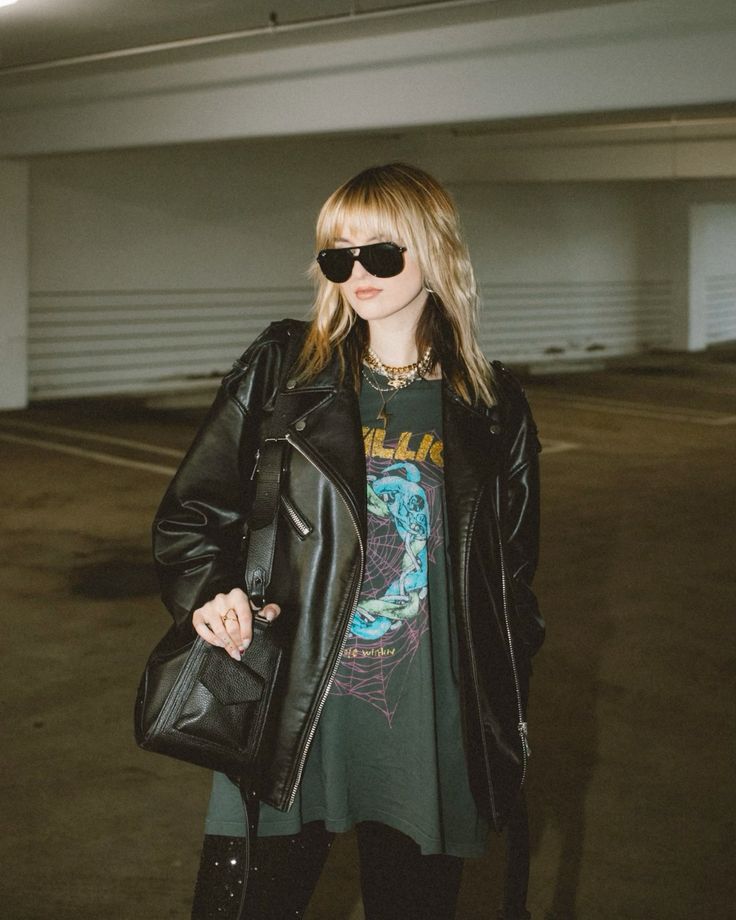 Person in stylish leather jacket and graphic tee, wearing sunglasses, posing in a dimly lit parking garage.