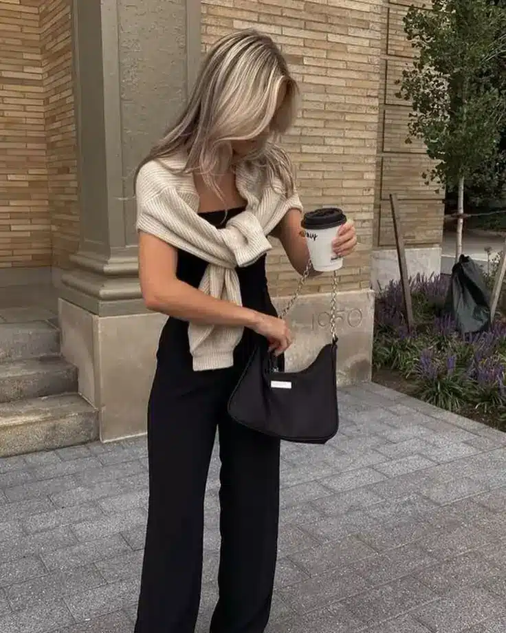 Woman in a stylish outfit holding a coffee cup, wearing a sweater, black attire, and a handbag, standing outside.