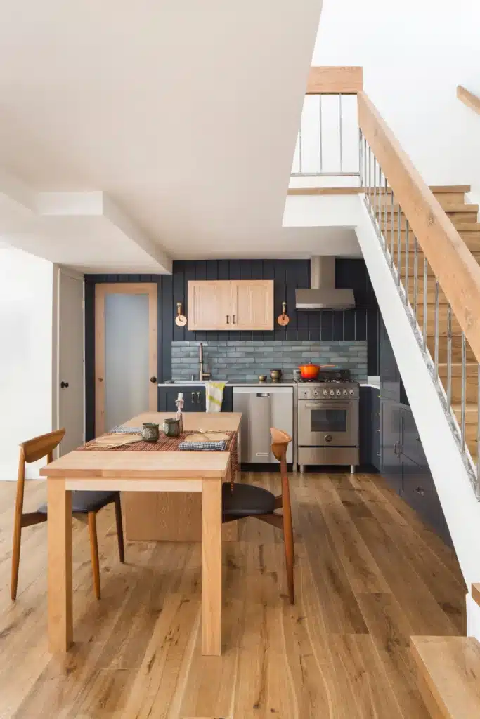 Modern kitchen with wooden dining table, blue cabinets, tiled backsplash, and staircase. Cozy, stylish interior design.