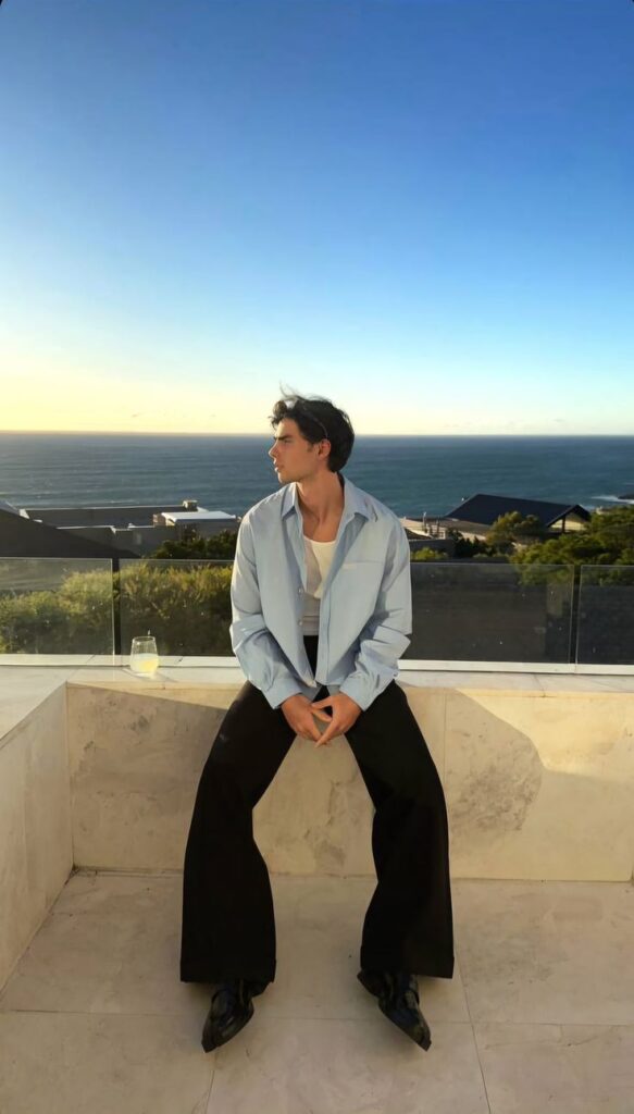 Man in light blue shirt sitting outdoors, gazing at ocean view during sunset.