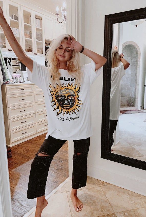 Person posing in casual outfit with graphic tee and ripped jeans, standing in a bright, stylish room with mirror reflection.