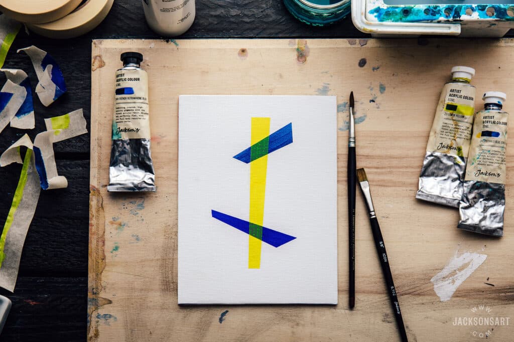 Abstract painting with blue and yellow stripes on canvas, surrounded by paint tubes and brushes on a wooden surface.