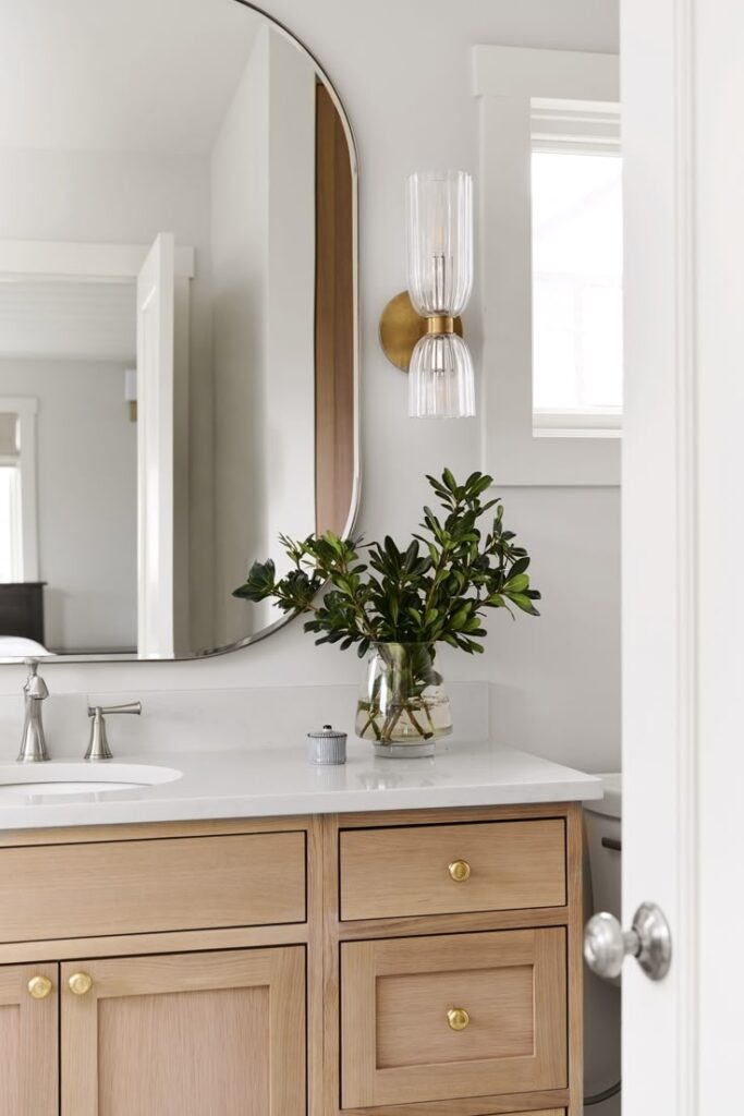 Modern bathroom vanity with oval mirror, wooden cabinetry, and vase with greenery on white countertop.