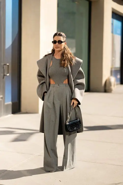 Stylish woman in gray outfit and sunglasses, walking on city street, carrying a chic black handbag.
