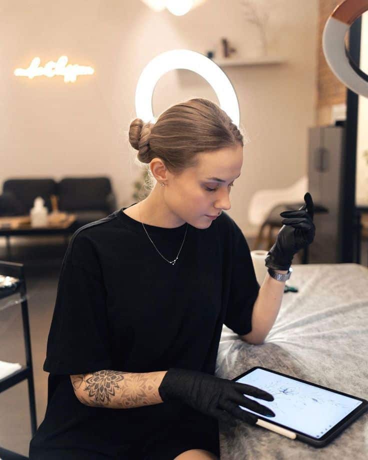 Tattoo artist working on digital design with tablet in studio, wearing gloves and surrounded by creative decor.