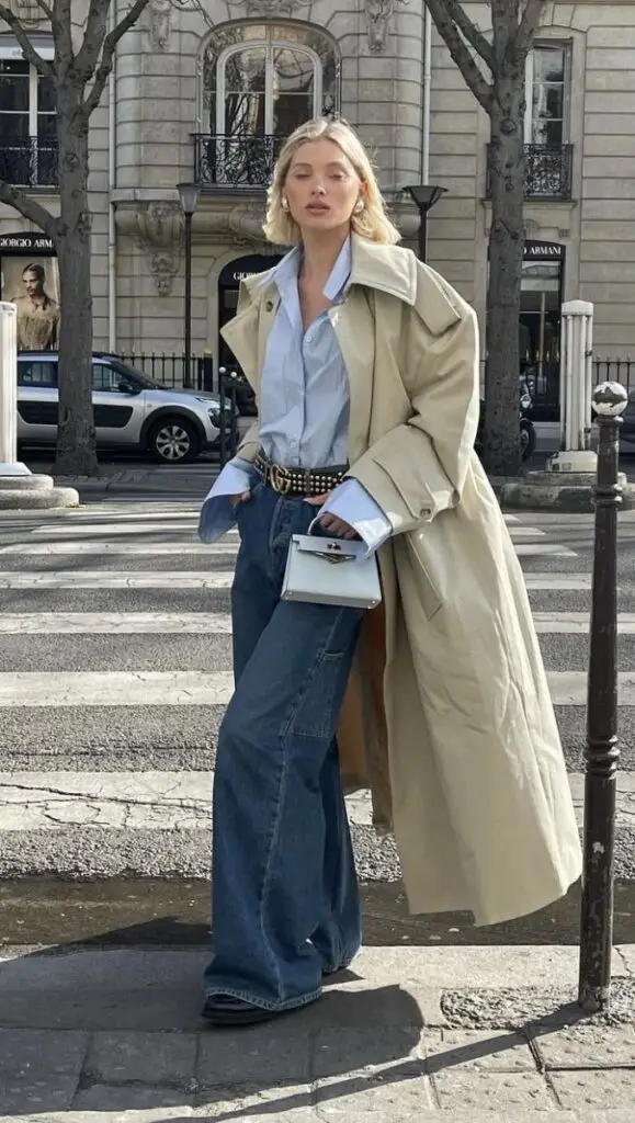 Fashionable woman in long beige coat, denim jeans, and light blue shirt, standing on a city street with small white purse.