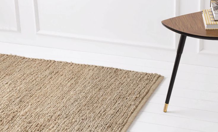 Beige textured rug on white wooden floor next to a modern table with books, enhancing minimalist interior design.