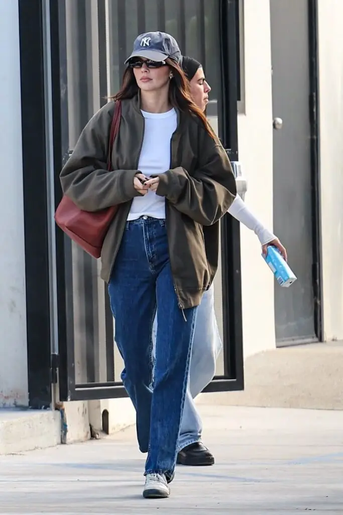 Casually dressed woman in jeans and cap, holding phone, walking outdoors, accompanied by another person with a drink.